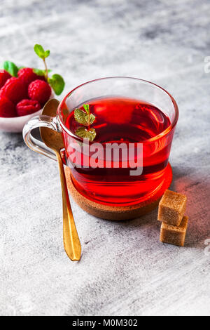 Heiße Himbeer Tee in transparenten Schale auf einem Tisch aus Stein. Frische Beeren, Würfel von Rohrzucker und eine Tüte Kaffee Stockfoto