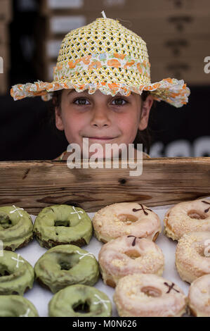 Junge Mädchen mit einem Schrulligen Lächeln beobachten Grün artisan Donuts ziemlich eng und trägt einen Strohhut auf einem Straßenfest in Marlybone London UK Stockfoto