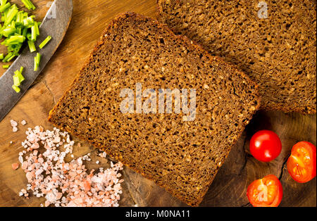 Schwarze Scheiben Vollkornbrot auf Holzuntergrund mit Schnittlauch, Tomaten, Himalaya-salz und alte Messer, flatlay, Ansicht von oben. Stockfoto