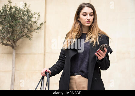 Junge Frau auf Handy. stehend gegen blonde Sandsteinmauer mit Oliven am ​Rear. lange dunkle Haare mit goldenen Boden Töne Stockfoto