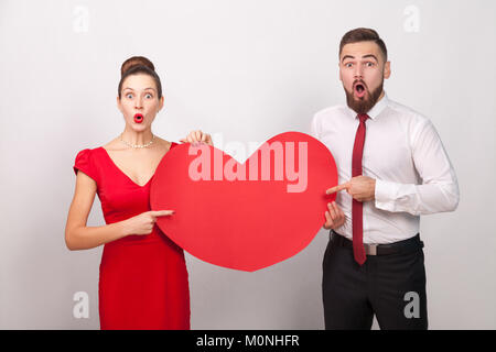 Wow! Große Liebe. Fragte sich paar Pointing Finger am Roten gehört. Innen-, Studio shot, auf grauem Hintergrund Stockfoto