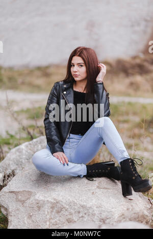 Attraktive schlanke junge Frau auf einem Felsen in einer trendigen schwarze Lederjacke und Spitze gekleidet sitzen - bis Stiefel nachdenklich auf die Kamera Stockfoto