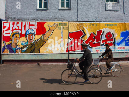 Die nordkoreanischen Männer Reiten Fahrräder vor der Propaganda Plakate in Hungnam Stickstoff-dünger Pflanze, Provinzen Süd-Hamgyong Provinz, Hamhung, Nordkorea Stockfoto