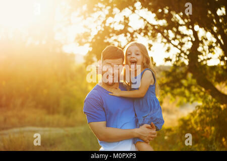 Der Vater hält die Tochter in die Arme. Urlaub für die ganze Familie. Sie Zunge. Ein Spaziergang im Stadtpark bei Sonnenuntergang. Stockfoto