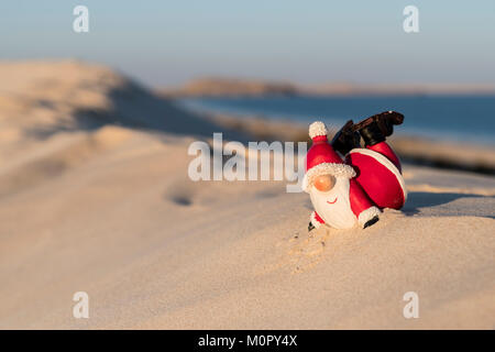 Die Figur des Weihnachtsmanns führt akrobatische Bewegungen an einem Strand aus, hinter dem sich der Ozean ausdehnt und festliche Stimmung mit einer lustigen Kulisse am Meer verbindet Stockfoto