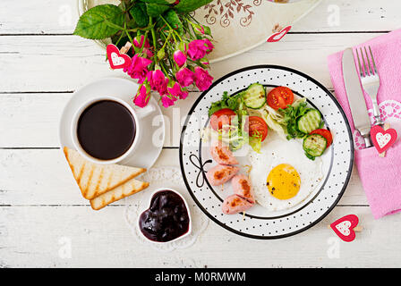 Frühstück am Valentinstag - Spiegelei in Herzform, Toast, Würstchen und frischem Gemüse. Tasse Kaffee. Englisches Frühstück. Ansicht von oben Stockfoto