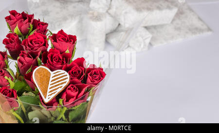 Blumenstrauß aus roten Rosen und süsses Herz für gutes Glück. Dekorative Blüten mit weißen Geschenke im Hintergrund. Feier, Geburtstag, Jubiläum, Valentinstag. Stockfoto