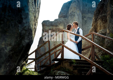 Romantische sensible Porträt der Jungvermählten halten sich an den Händen und gehen auf die hölzerne Treppe zwischen Felsen während des Sonnenuntergangs zu küssen. Stockfoto