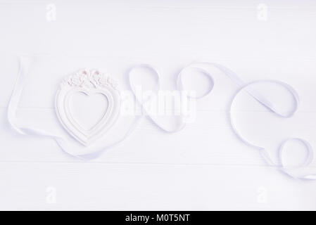 Ansicht von oben Liebe Wort Schriftzug mit weißem Satinband und Bilderrahmen in Form von Herzen auf dem weißen Holz- Hintergrund geschrieben. Flach. Liebe Ausdruck. Kopieren Sie Platz. Stockfoto