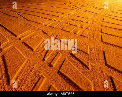 Die Spuren von Autoreifen sind im Schnee unter Natriumlicht sichtbar und halten einen Moment der kalten Winterlandschaft mit den markanten Markierungen auf dem Schnee fest. Stockfoto