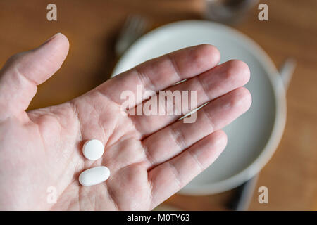 Nahaufnahme von einem weißen männlichen Hand zwei weiße Pillen in der Handfläche über einen verschwommenen Hintergrund zeigt einen Esstisch mit Teller, Besteck und Glas. Stockfoto