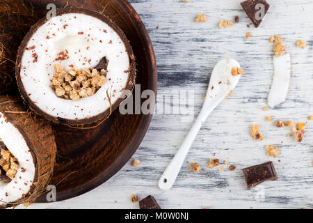 Gesundes Frühstück in Coconut Bug auf weißem Hintergrund. Joghurt in Kokosnuss Schale mit Kokosraspeln, Schokolade und Müsli. Ansicht von oben, flach, Overhead Stockfoto