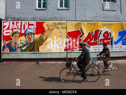 Die nordkoreanischen Männer Reiten Fahrräder vor der Propaganda Plakate in Hungnam Stickstoff-dünger Pflanze, Provinzen Süd-Hamgyong Provinz, Hamhung, Nordkorea Stockfoto