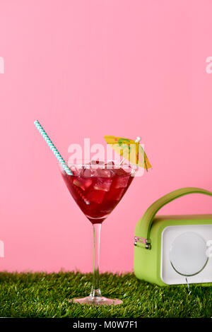 Ein Cocktail Glas mit einem roten Getränk garniert mit einem Regenschirm und eine retro Grün Radio auf dem Rasen, gegen einen rosa Hintergrund mit einem leeren Raum Stockfoto