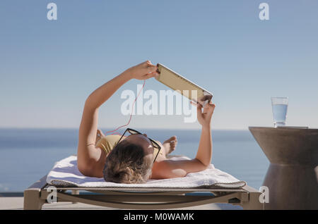 Frau sonnenbaden, mit digitalen Tablette auf Lounge Chair auf der sonnigen Terrasse mit Blick auf den Ozean Stockfoto