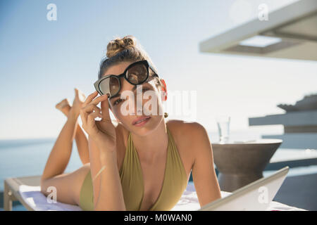 Portrait ernst, selbstbewusste Frau im Badeanzug und Sonnenbrille Sonnenbaden mit digitalen Tablet an sonnigen Luxus Terrasse Stockfoto