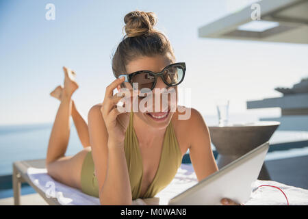 Lächelnde Frau im Badeanzug und Sonnenbrille mit digitalen Tablet, Sonnenbaden an Lounge Chair auf der sonnigen Terrasse Stockfoto