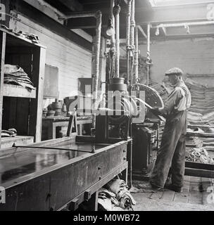 1950er Jahre, historische Bild eines männlichen Arbeitnehmers in einer Reifenfabrik, 'Lage', die Schichten der Gewebe, in der Regel LWL-Kabel, das in einer Lösung aus Gummi beschichtet sind - der schwarze Gummibärchen Compound in der langen Tank gesehen -, die das Skelett der Reifen zu machen, damit es flexibel, aber nicht elastisch, England, UK. Stockfoto