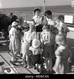 Mitte der 1950er-Jahre, historische, an Bord eines Dampfers Union-Castle mid-ocean gehen für die Kap in Südafrika, Post, Fracht und Passagieren, eine Gruppe von Kindern sammeln auf dem Deck um ein grammophon Player, mit einer Pflegekraft des Schiffes und eine andere Dame überwachen. Stockfoto