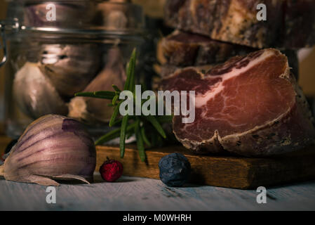 Horizontale Foto von Schweinefleisch von Wildschweinen, die getrocknet und geräuchert. Die Stücke sind gestapelt. Italienisch und Toskana traditionelle Lebensmittel. Knoblauch, Beeren Stockfoto