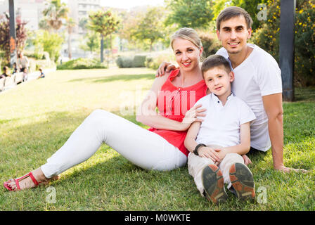 Junge fröhliche amerikanische Elternteil mit Junge im Teenager-Alter sitzen auf grünen Gras im Park Stockfoto
