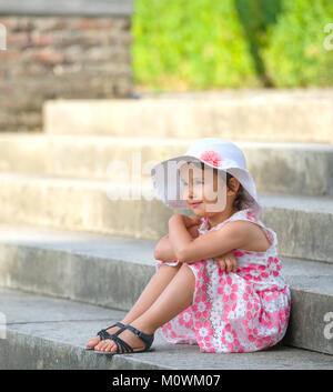 Adorable kleine Mädchen mit weißen Hut sitzt auf der Treppe, an warmen und sonnigen Sommer Tag Stockfoto