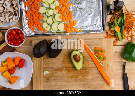 Vorbereitung verschiedene Gemüse kochen mit dem halbierte Avocado pear auf einer hölzernen Schneidebrett umgeben mit gewürfelten Karotten, Tomaten, mushroo Stockfoto