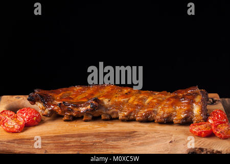 Schweinerippchen in Barbecue Sauce und Honig gerösteten Tomaten auf einem Holzbrett. Ein toller Snack zum Bier auf einem schwarzen Hintergrund. Mit kopieren. Stockfoto