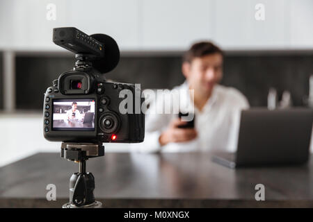 Junger Mann nimmt sein Video Blog Episode über die neuen technischen Geräte während am Küchentisch sitzen und mit leerer Bildschirm der mobilen Pho konzentriert Stockfoto