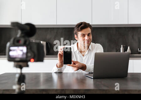 Junger Mann nimmt sein Video Blog Episode über die neuen technischen Geräte während am Küchentisch sitzen und mit leerer Bildschirm der mobilen Pho konzentriert Stockfoto