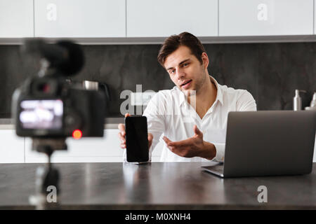 Hübscher junger Mann nimmt sein Video Blog Episode über die neuen technischen Geräte während am Küchentisch sitzen und mit leerer Bildschirm des Mobiltelefons Stockfoto