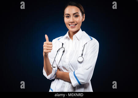Junge positive Frau Doktor mit Stethoskop mit Daumen und lächelnd isoliert Stockfoto
