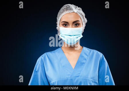 Junger Arzt in der medizinischen Maske und Kappe tragen blaue Uniform und suchen Kamera über Schwarz isoliert Stockfoto