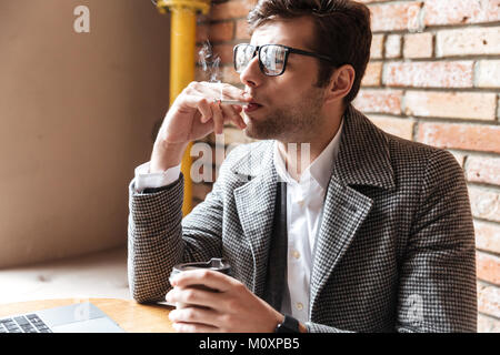 Seitenansicht der junge Geschäftsmann in Brillen saßen am Tisch im Cafe und Rauchen Zigarette Stockfoto
