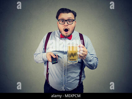 Mollig Mann holding Becher Bier, während der TV-Programmwiedergabe und Suchen erstaunt. Stockfoto