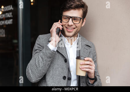 Gerne business Mann in Brillen und Mantel sprechen von Smartphone bei einer Tasse Kaffee in der Hand???????? Stockfoto