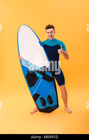 Volle Länge Portrait von glücklich aussehenden Surfer angezeigt "Daumen hoch" Geste beim Stehen und halten ein Surfbrett, über Gelb Hintergrund isoliert Stockfoto