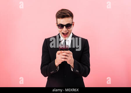 Überrascht lächelnd Mann in offiziellen Anzug und Sonnenbrille mit Smartphone über rosa isoliert Stockfoto