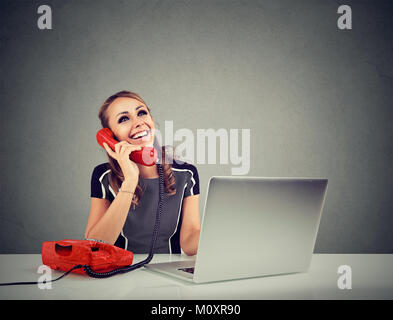 Junge fröhliche Frau in Telefongespräch während am Schreibtisch arbeiten am Laptop sitzen. Stockfoto