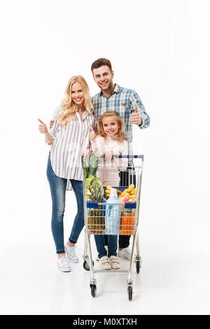 In voller Länge Porträt einer glücklichen Familie mit einem Einkaufswagen voller Lebensmittel und zeigt den Daumen oben-Geste auf weißem Hintergrund Stockfoto