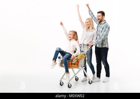 In voller Länge Porträt einer fröhlichen Familie wandern mit einem Einkaufswagen voller Lebensmittel auf weißem Hintergrund, kleines Mädchen im Auto sitzen Stockfoto