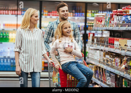 Kleines Mädchen sitzt auf einem Warenkorb und Auswahl von Candy mit ihren Eltern im Supermarkt begeistert Stockfoto