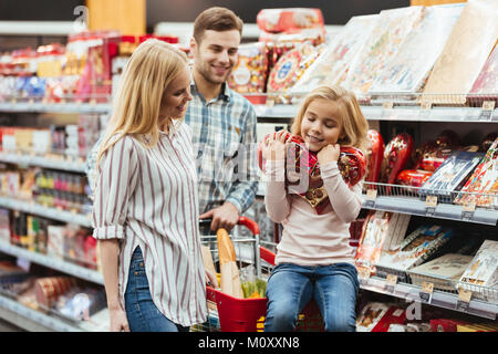 Lächelnde Mädchen sitzt auf einem Warenkorb und Auswahl von Candy mit ihren Eltern im Supermarkt Stockfoto