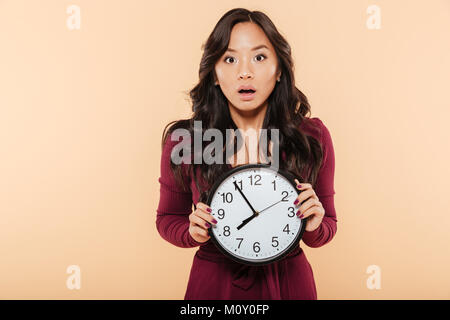 Junge asiatische Frau mit dem lockigen langen Haare, die Uhr zeigt fast 8, Ende, oder Fehlt etwas über Pfirsich Hintergrund Stockfoto