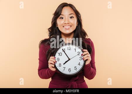 Glückliche Gefühle der asiatische Frau mit dem lockigen langen Haare, die Uhr zeigt fast 8, warten auf etwas Angenehmes über Pfirsich Hintergrund Stockfoto