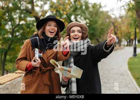 Porträt von zwei fröhliche Mädchen im Herbst Kleidung holding Stadtplan beim stehen draußen und Pointing Finger weg gekleidet Stockfoto
