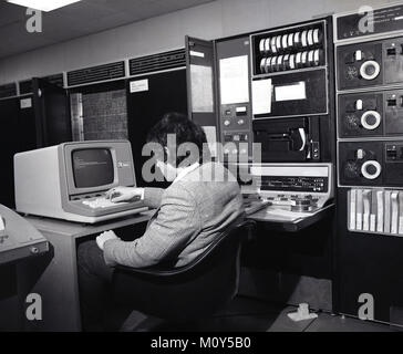 1970 s, historische Bild von einem Mann in einem EDV-Raum mit Banken von elektronischen Geräten neben ihm, an einem kleinen Arbeitsplatz mit einem Computer Terminal mit Bildschirm, England, Großbritannien sitzen. Stockfoto