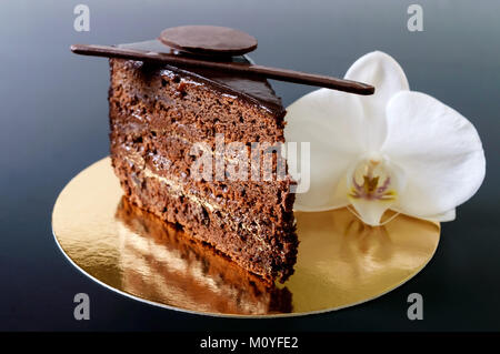 Ein Stück Schokoladenkuchen mit Schokoladencreme auf einem goldenen Serviette mit einer weißen Orchidee auf schwarzem Hintergrund. Stockfoto