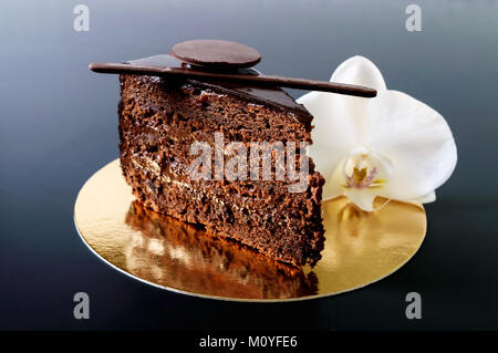 Ein Stück Schokoladenkuchen mit Schokoladencreme auf einem goldenen Serviette mit einer weißen Orchidee auf schwarzem Hintergrund. Stockfoto