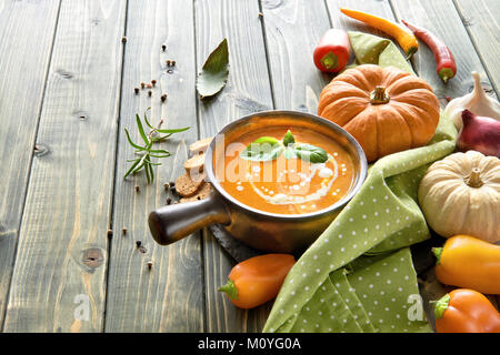 Pikante Kürbiscremesuppe mit gebratenen Paprika und Chili in einer dunklen Keramik Topf auf dem Holztisch, gewürzt mit Sahne und Basilikum. Stockfoto